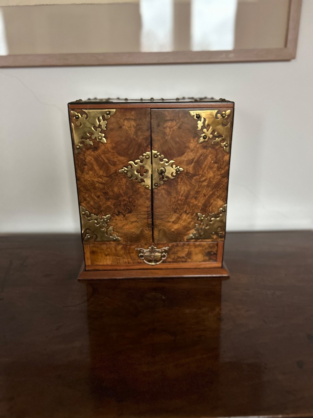 A Victorian burr walnut cigar cabinet featuring two doors adorned with pierced brass corner brackets and a central lock mechanism above a lower drawer. View VI