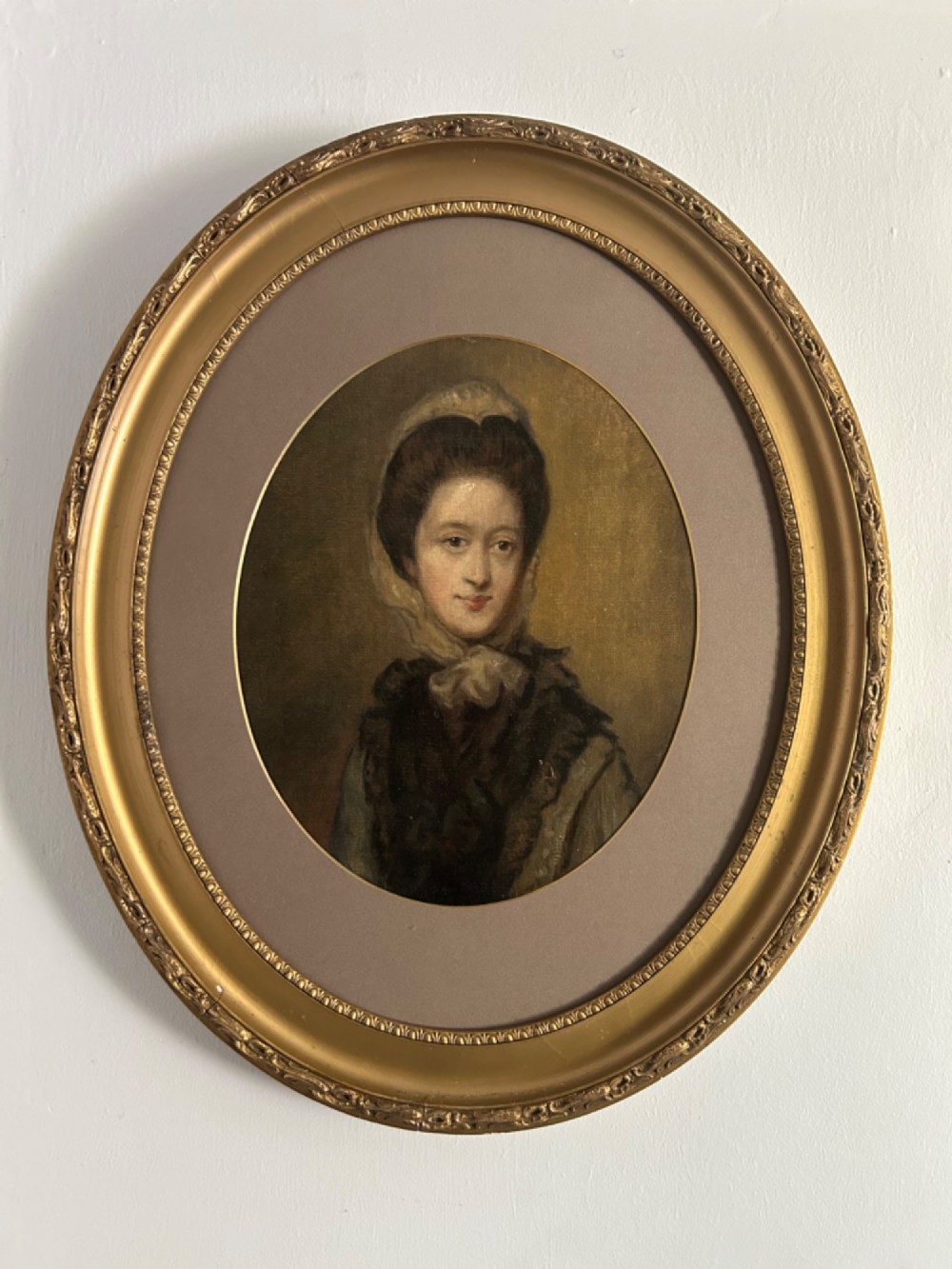 A late eighteenth-century oval oil portrait depicting a woman wearing a bonnet and dark mantle, presented inside a gilded frame.
