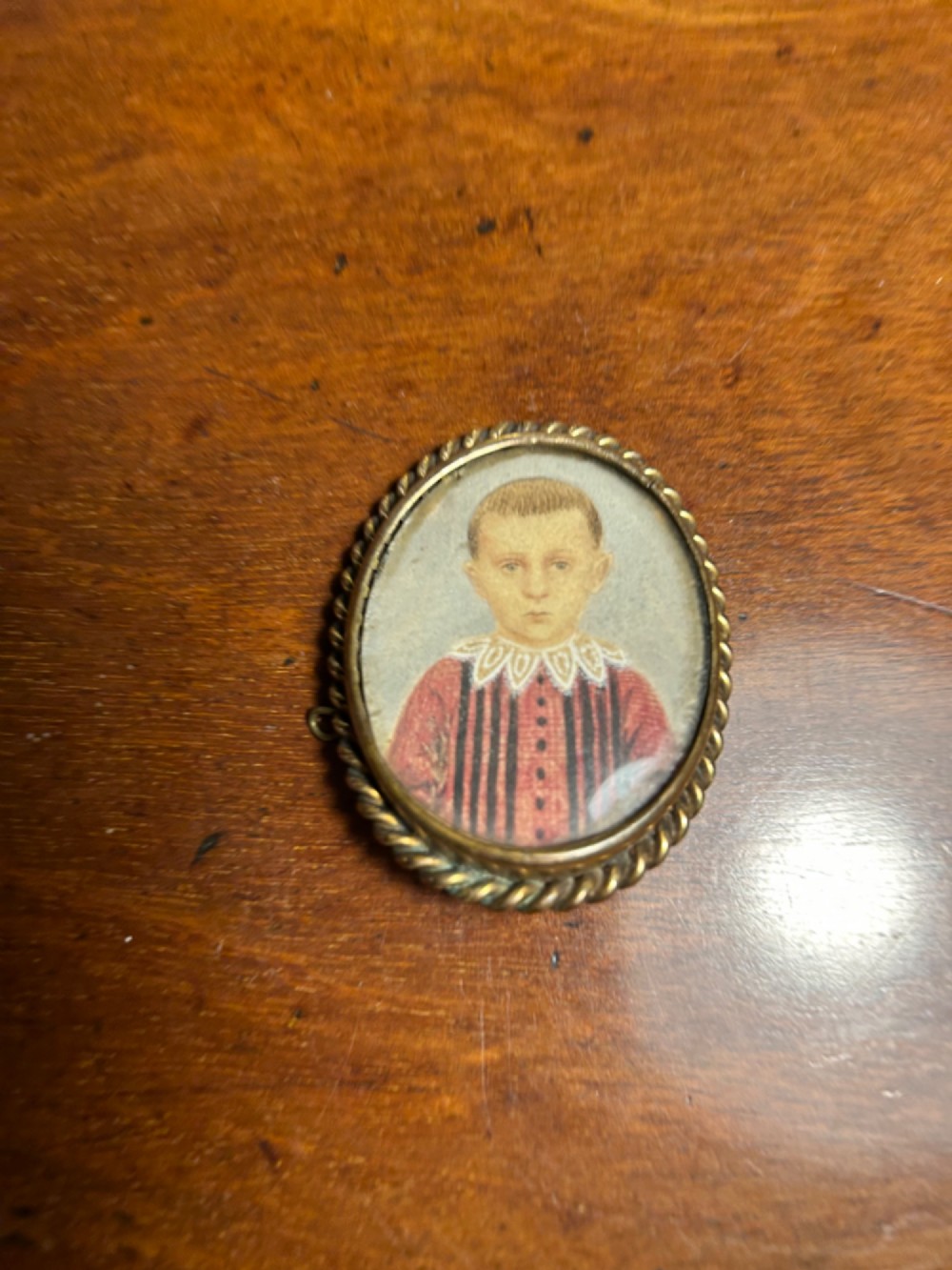 An oval Victorian portrait miniature depicting a young boy in a red striped garment set within a gold-toned rope-twist metal brooch frame. View III