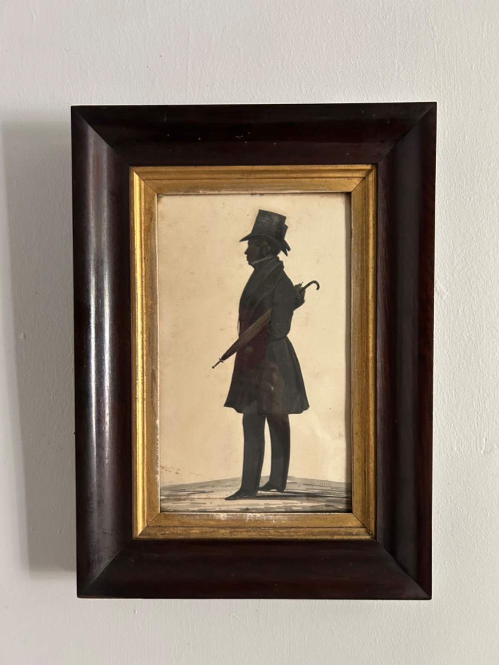 A nineteenth-century full-length silhouette portrait of a gentleman in a top hat holding an umbrella, presented in a gilded slip and a dark wood frame.