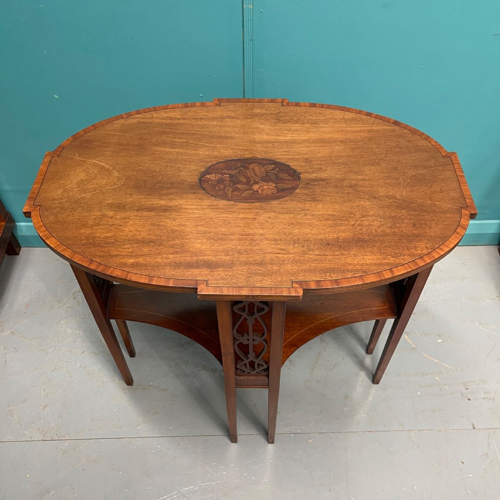 An Edwardian oval wooden side table featuring a central floral marquetry medallion, crossbanded border, pierced fretwork side aprons, and an under-shelf.