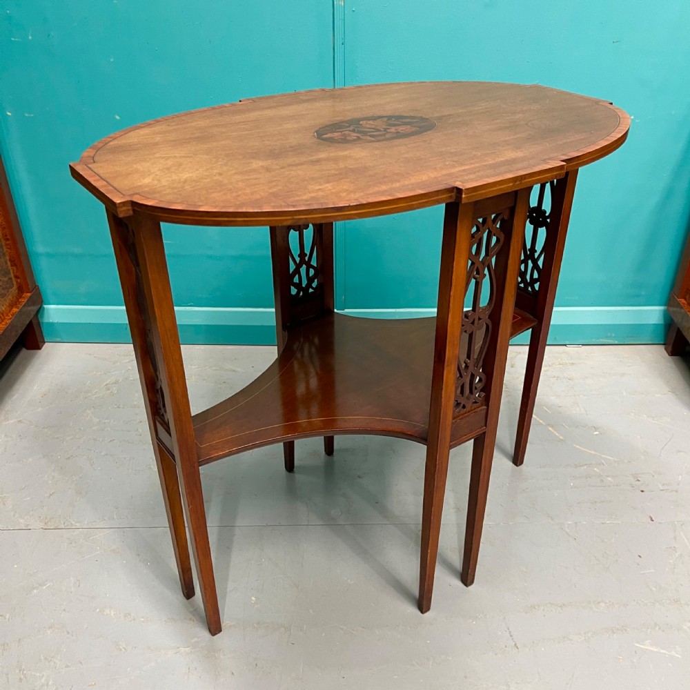 An Edwardian oval wooden side table featuring a central floral marquetry medallion, crossbanded border, pierced fretwork side aprons, and an under-shelf. View V