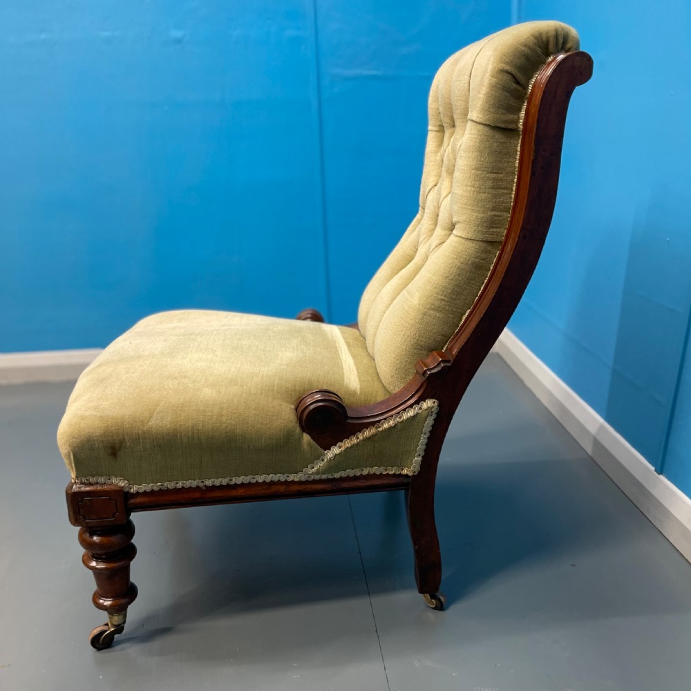 A William IV slipper chair upholstered in olive green velvet with deep button tufting, a scroll back, and turned mahogany front legs on brass castors. View IV