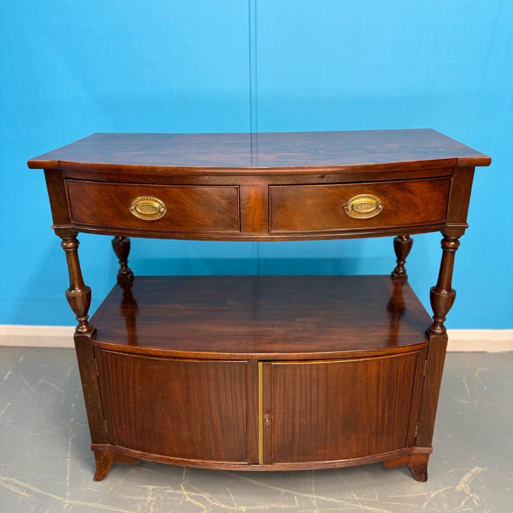 A dark mahogany Georgian bow front buffet cabinet featuring two upper drawers with oval brass handles, turned side columns, an open centre shelf, and a lower double-door cupboard.