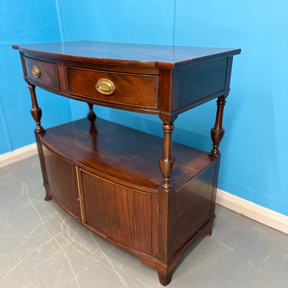 A dark mahogany Georgian bow front buffet cabinet featuring two upper drawers with oval brass handles, turned side columns, an open centre shelf, and a lower double-door cupboard. View II