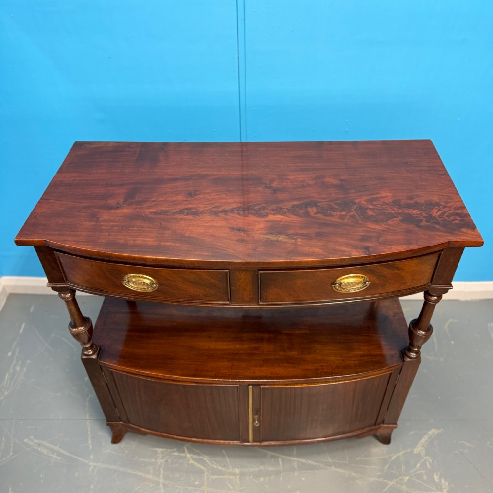 A dark mahogany Georgian bow front buffet cabinet featuring two upper drawers with oval brass handles, turned side columns, an open centre shelf, and a lower double-door cupboard. View III