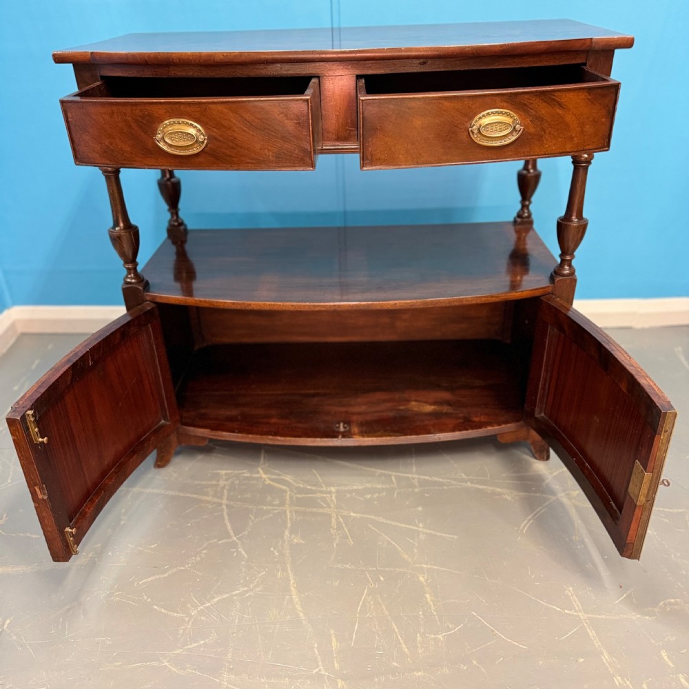 A dark mahogany Georgian bow front buffet cabinet featuring two upper drawers with oval brass handles, turned side columns, an open centre shelf, and a lower double-door cupboard. View IV