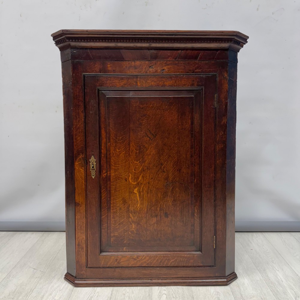 An eighteenth-century Georgian dark oak corner cupboard featuring a single raised panel door with a decorative brass keyhole escutcheon and moulded cornice.