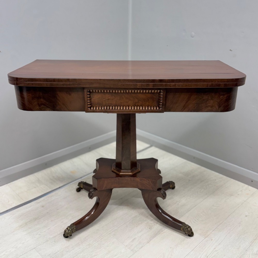 A Late Georgian dark mahogany fold-over side table featuring a beaded frieze, faceted pedestal base, and quadruped sabre legs with brass paw castors.