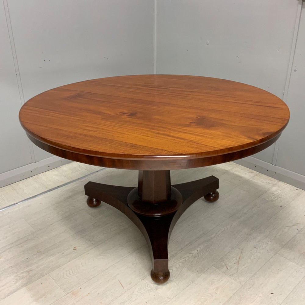 A circular Regency mahogany centre table featuring a smooth timber top supported on a central pedestal and a concave triform base with ball feet.