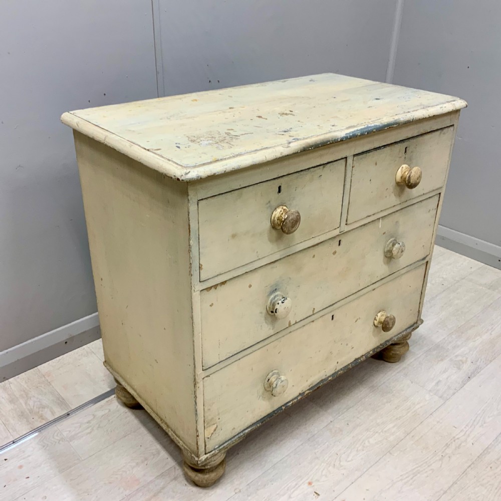 A 19th-century painted pine chest of drawers featuring two short over two long drawers, turned wooden knobs, turned bun feet, and a distressed cream painted finish.
