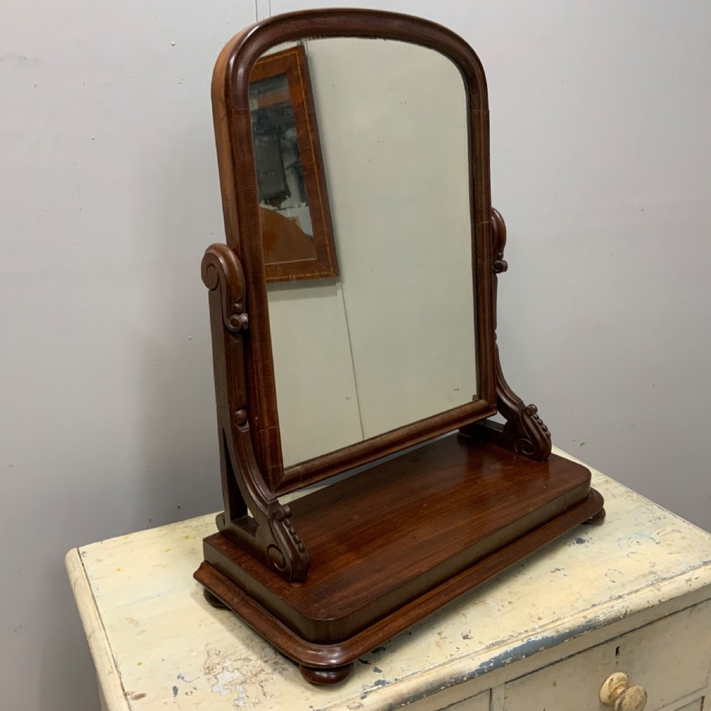 An antique Victorian mahogany toilet mirror featuring an arched glass frame supported by carved scroll uprights on a solid wooden plinth base.