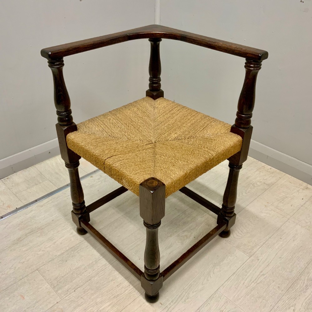 A dark oak corner chair featuring turned legs, a continuous curved armrest, and a hand-woven natural rush seat.