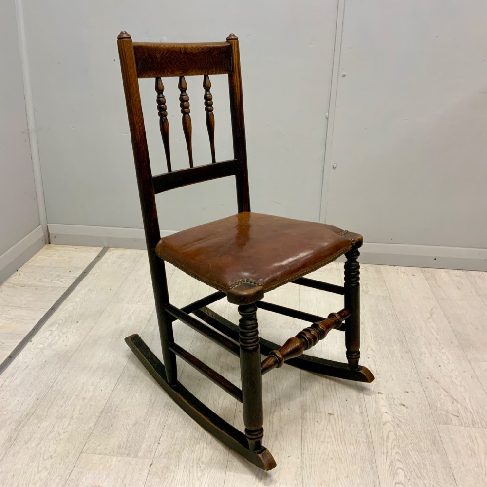 A mid-19th century dark oak rocking chair featuring a spindle backrest, a brown studded leather seat, and turned wooden front legs.