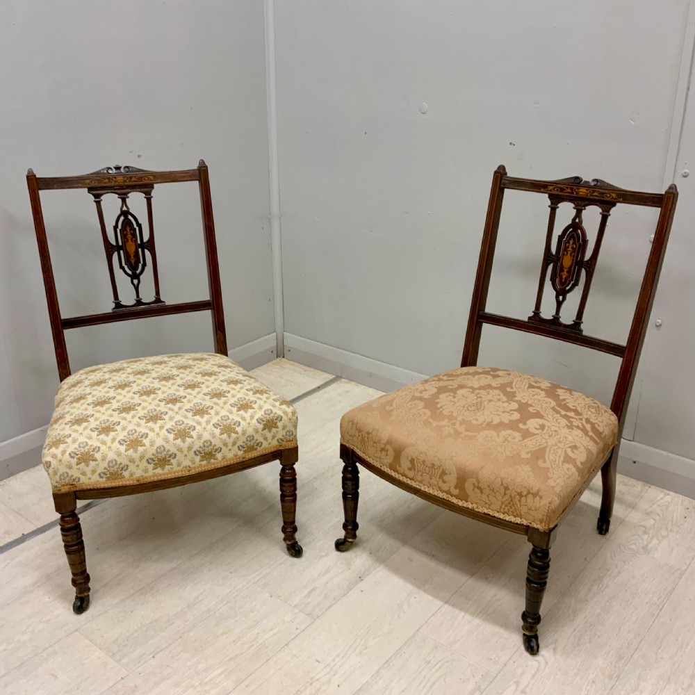 A pair of Edwardian inlaid mahogany nursing chairs featuring splats decorated with marquetry, padded upholstered seats, and turned front legs on castors.