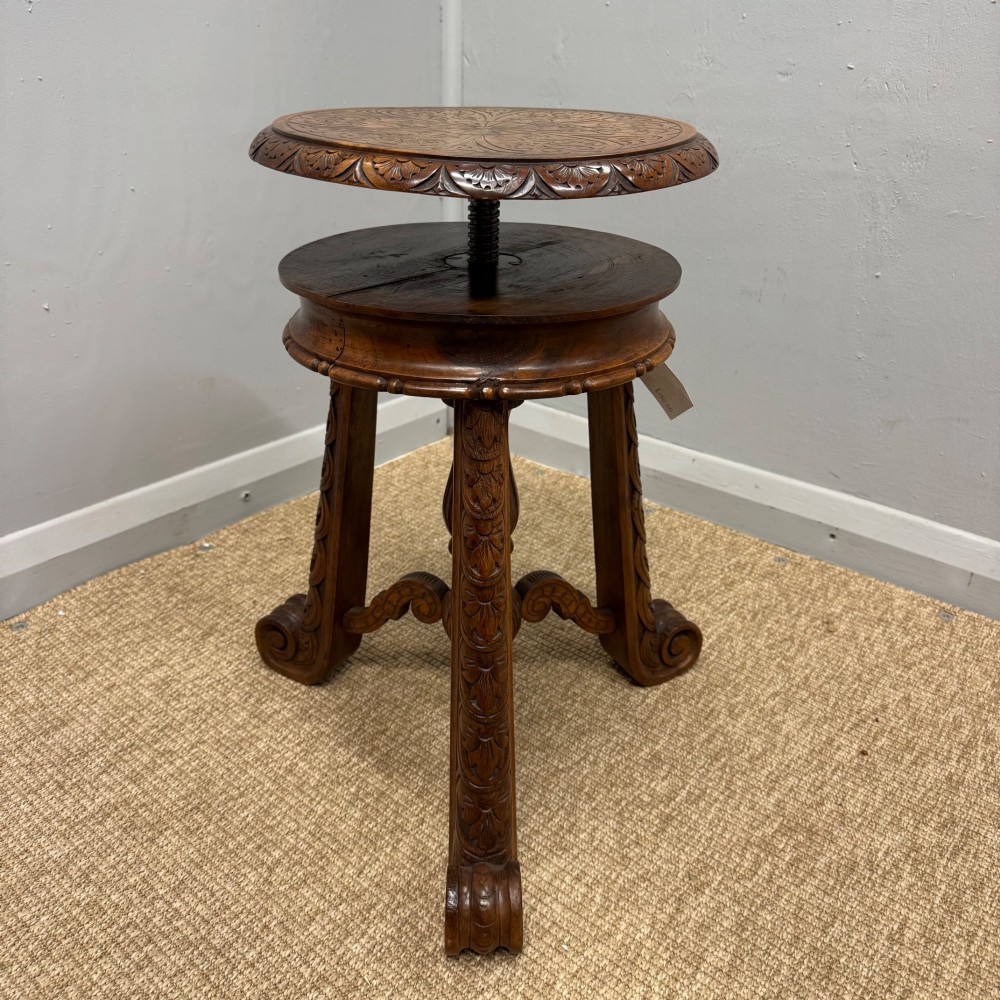 A carved dark hardwood revolving piano stool featuring a circular medallion seat, supported by three carved scroll legs and a central turned column. View II
