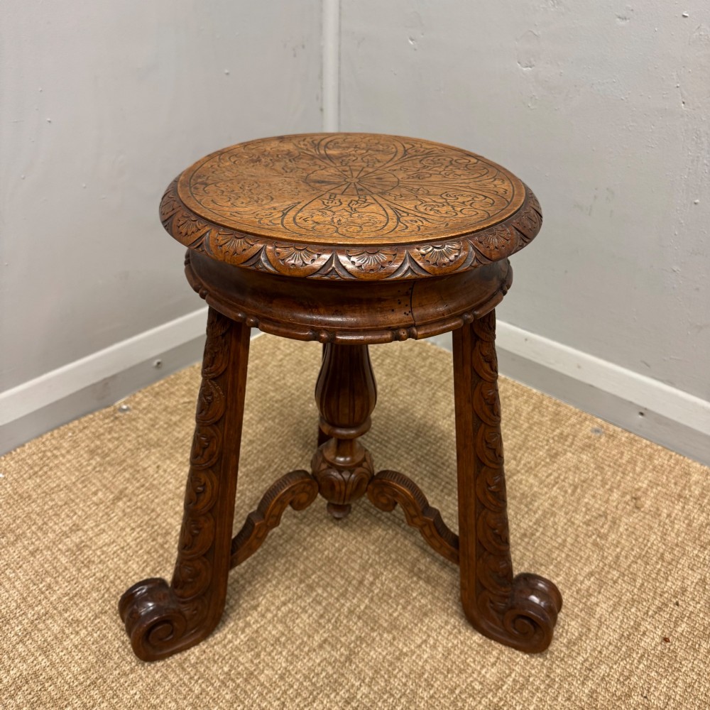 A carved dark hardwood revolving piano stool featuring a circular medallion seat, supported by three carved scroll legs and a central turned column. View IV