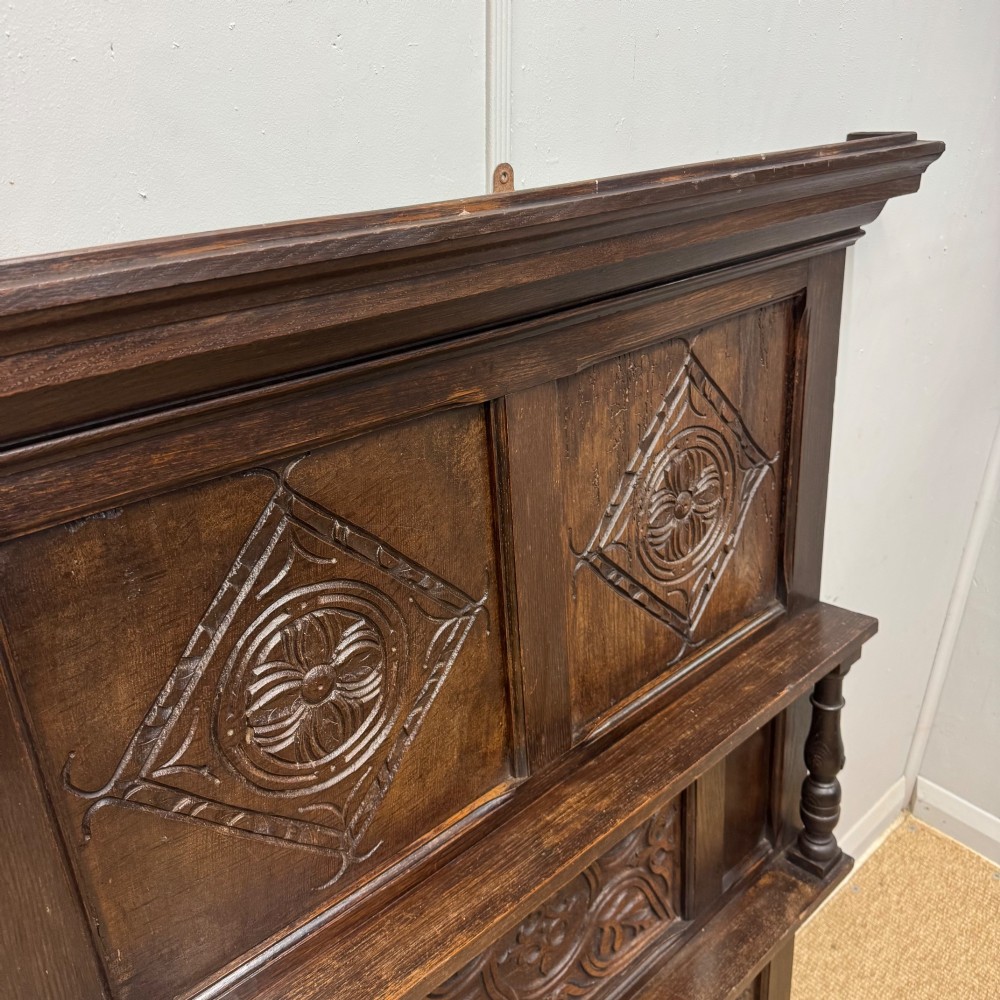 A 19th-century dark carved oak overmantel panel featuring carved geometric lozenges, a central foliate panel, turned spindle supports, and two display shelves. View II