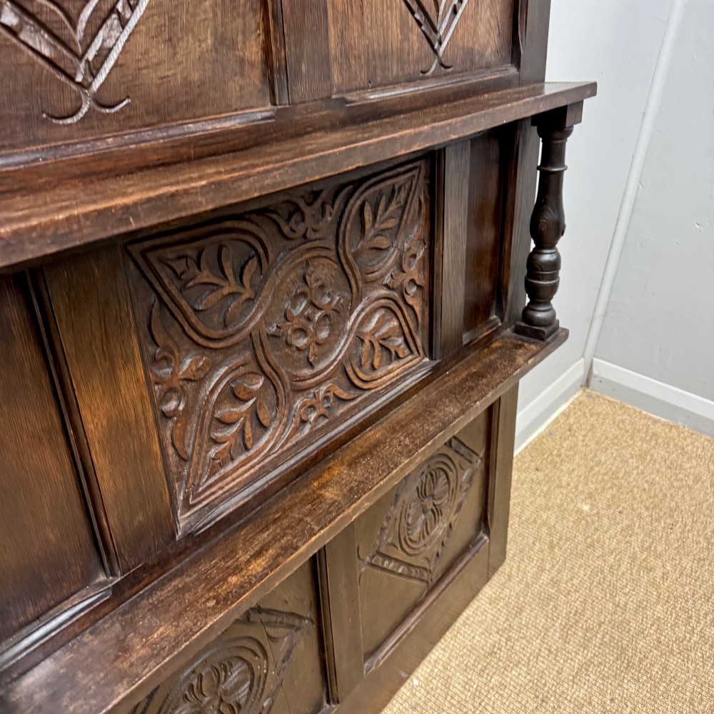 A 19th-century dark carved oak overmantel panel featuring carved geometric lozenges, a central foliate panel, turned spindle supports, and two display shelves. View IV