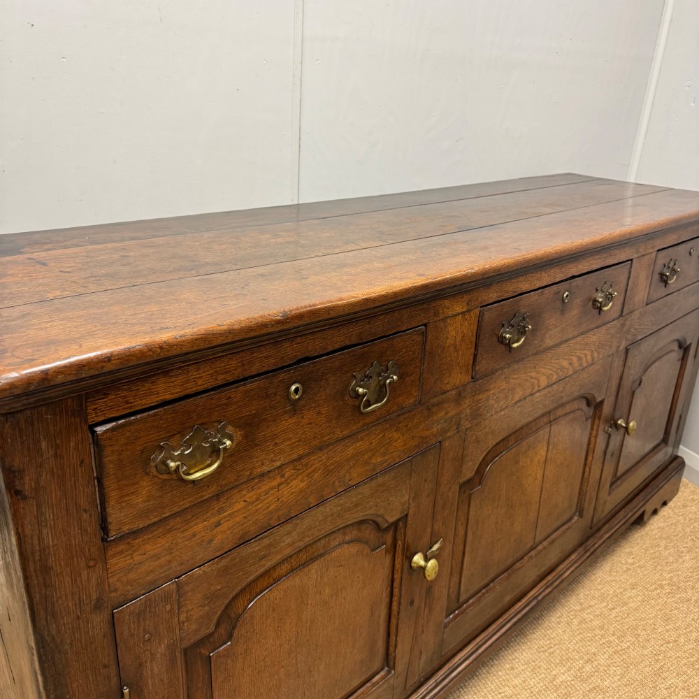 An 18th-century Georgian oak dresser base featuring three top drawers with brass drop handles above three cupboard doors with fielded arched panels. View VI