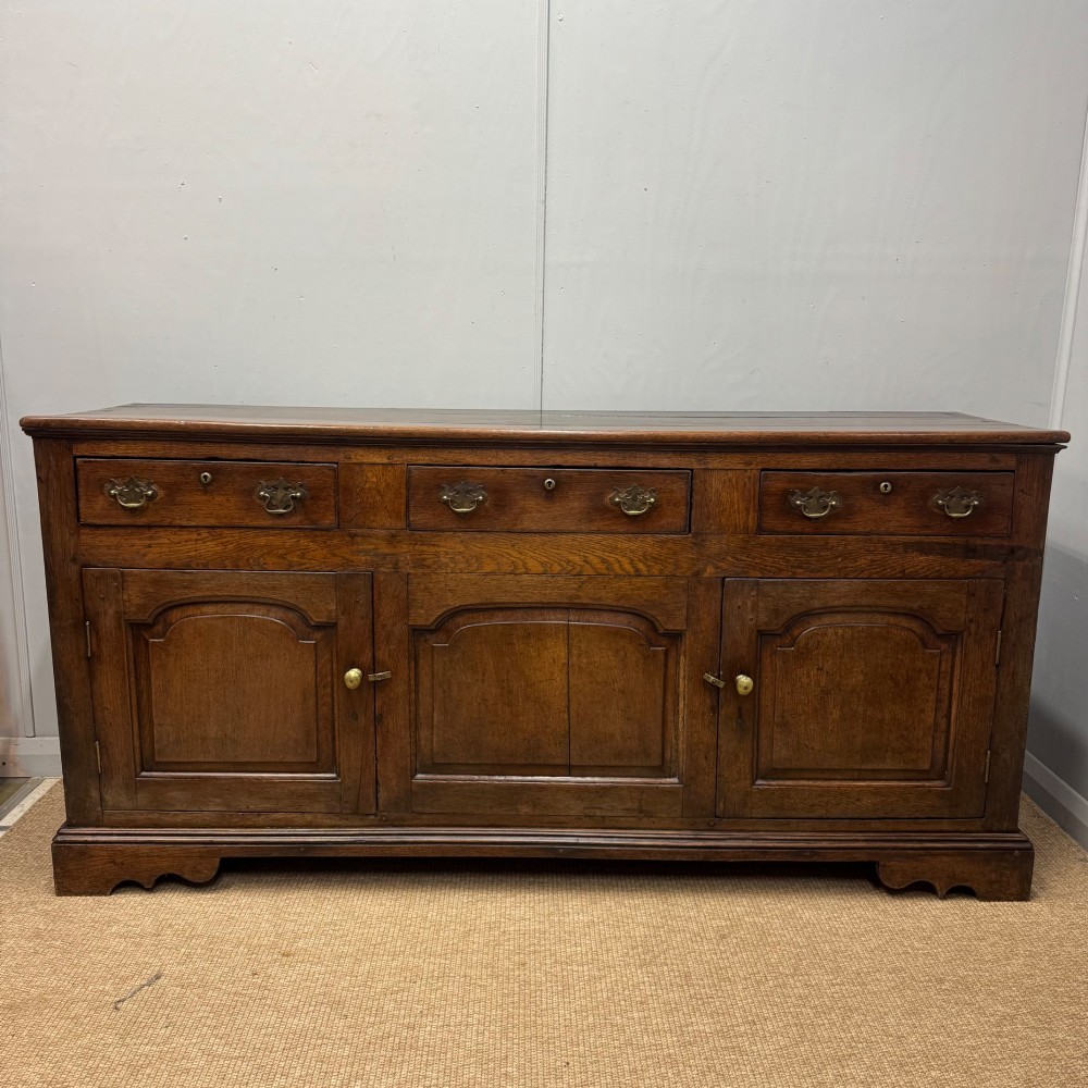 An 18th-century Georgian oak dresser base featuring three top drawers with brass drop handles above three cupboard doors with fielded arched panels. View IX