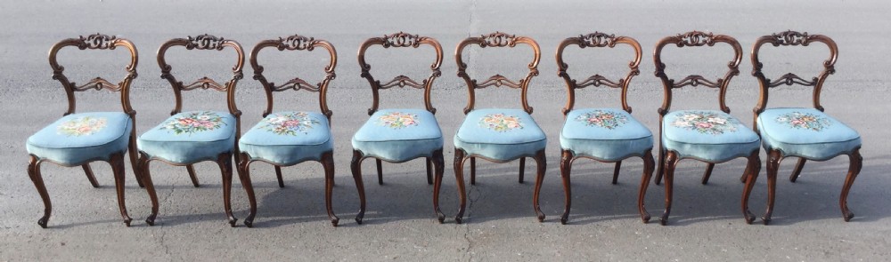 A set of eight Victorian walnut dining chairs featuring carved balloon backs, light blue velvet upholstered seats with central floral needlepoint designs, and cabriole front legs.