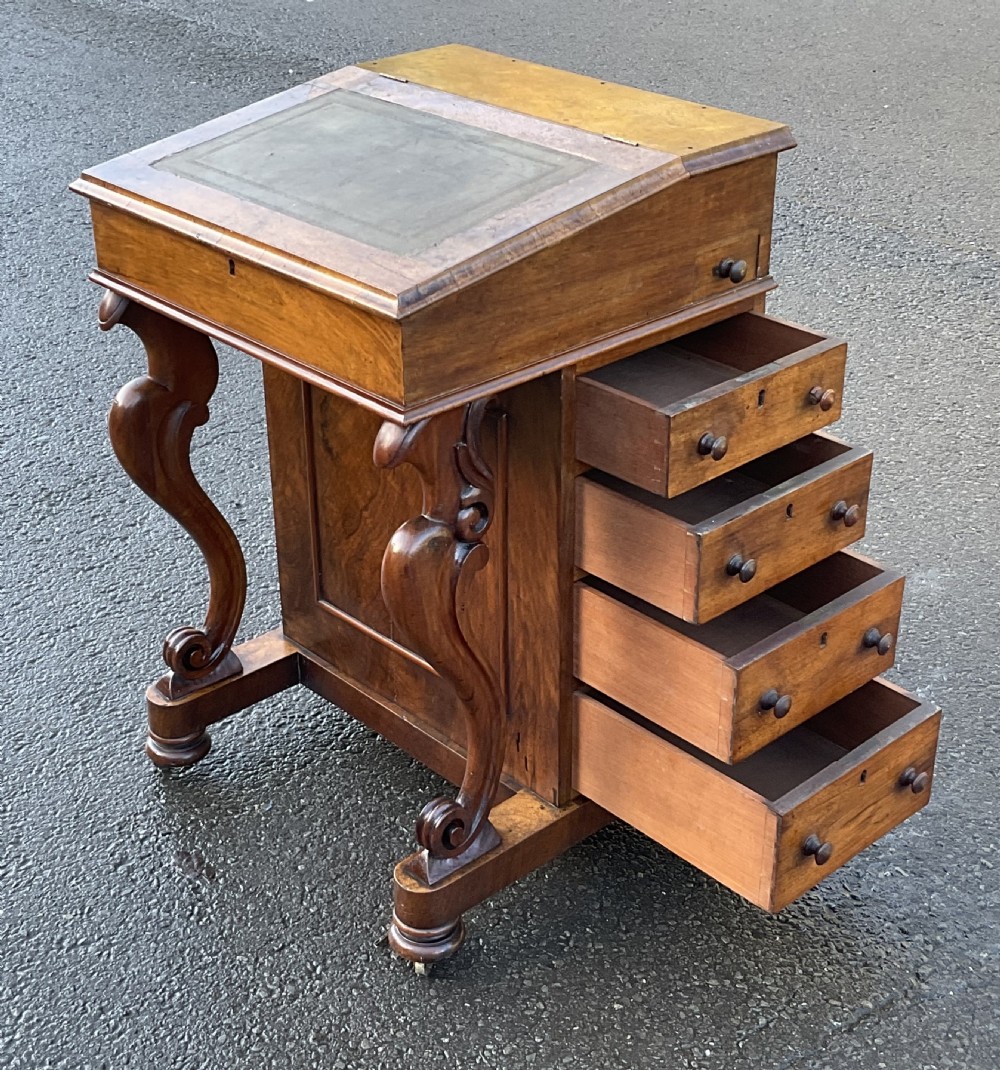 A Victorian walnut Davenport desk featuring a slanted writing surface with a green leather inset, side drawers with turned wooden knobs, and carved S-scroll front supports. View X