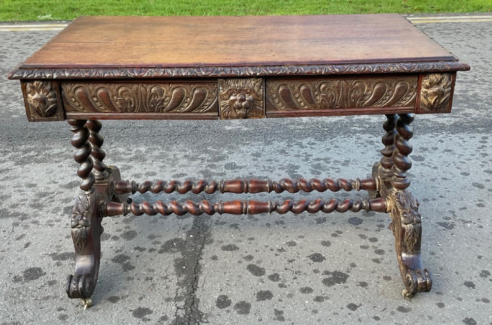 A Victorian dark oak writing table featuring a flat rectangular top, relief-carved frieze with mask motifs, barley twist legs, double stretchers, and brass castors.