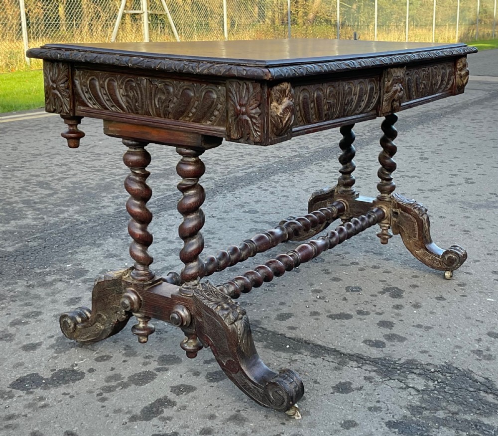 A Victorian dark oak writing table featuring a flat rectangular top, relief-carved frieze with mask motifs, barley twist legs, double stretchers, and brass castors. View II