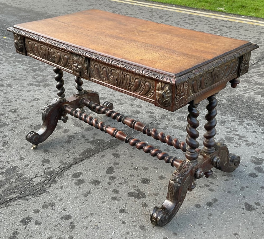 A Victorian dark oak writing table featuring a flat rectangular top, relief-carved frieze with mask motifs, barley twist legs, double stretchers, and brass castors. View III