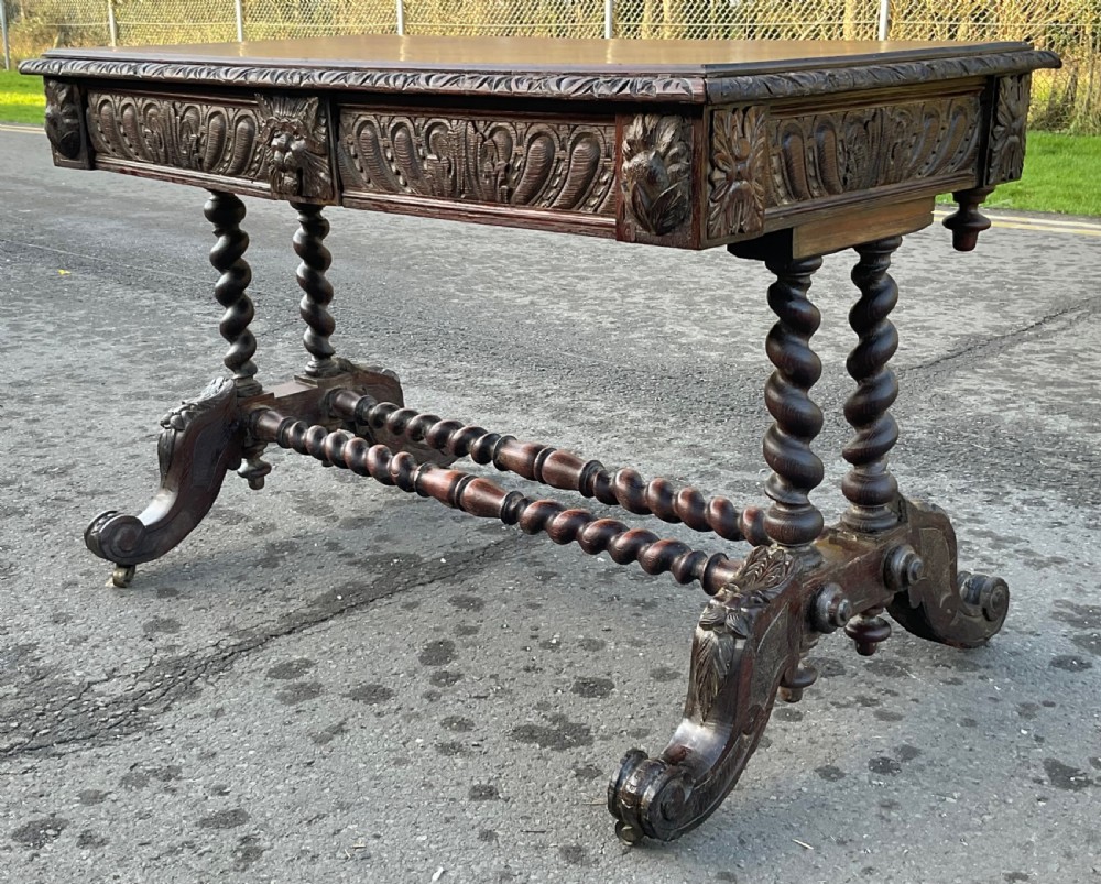 A Victorian dark oak writing table featuring a flat rectangular top, relief-carved frieze with mask motifs, barley twist legs, double stretchers, and brass castors. View V