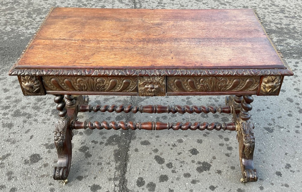 A Victorian dark oak writing table featuring a flat rectangular top, relief-carved frieze with mask motifs, barley twist legs, double stretchers, and brass castors. View VI
