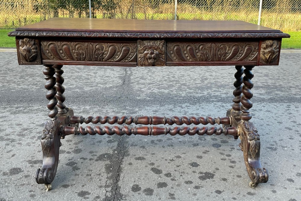 A Victorian dark oak writing table featuring a flat rectangular top, relief-carved frieze with mask motifs, barley twist legs, double stretchers, and brass castors. View VII