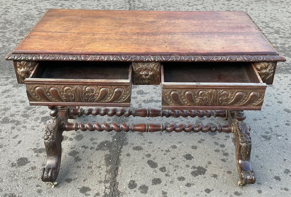 A Victorian dark oak writing table featuring a flat rectangular top, relief-carved frieze with mask motifs, barley twist legs, double stretchers, and brass castors. View IX