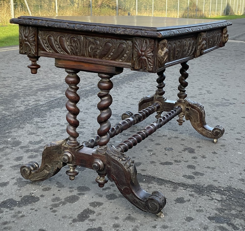 A Victorian dark oak writing table featuring a flat rectangular top, relief-carved frieze with mask motifs, barley twist legs, double stretchers, and brass castors. View X