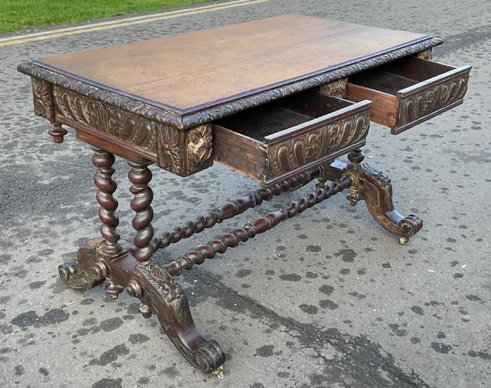 A Victorian dark oak writing table featuring a flat rectangular top, relief-carved frieze with mask motifs, barley twist legs, double stretchers, and brass castors. View XII