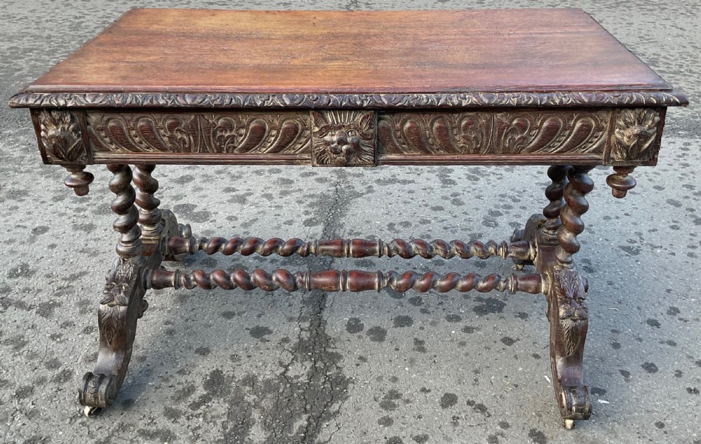A Victorian dark oak writing table featuring a flat rectangular top, relief-carved frieze with mask motifs, barley twist legs, double stretchers, and brass castors. View XIII