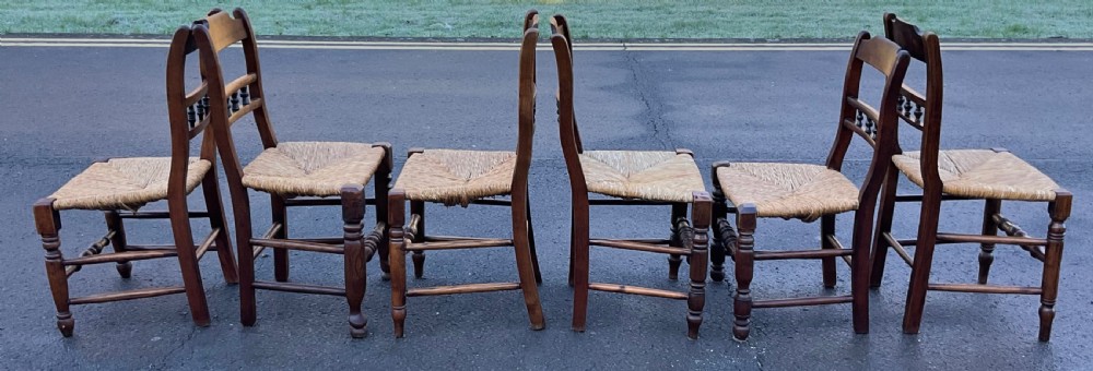 A set of six dark wooden elm dining chairs featuring hand-woven rush seats, turned front stretchers, and spindle back detail. View IV