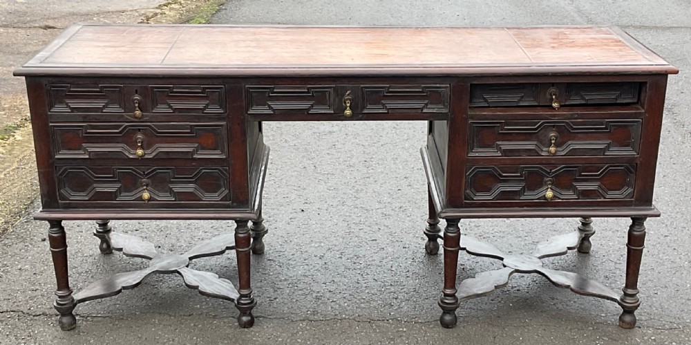 A large dark oak pedestal writing desk featuring geometric panelled drawer fronts, a leather inset writing surface, turned legs, and cross stretchers.