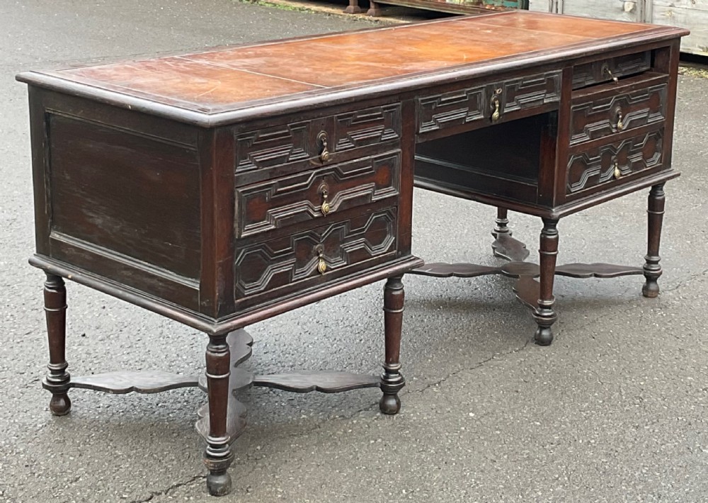 A large dark oak pedestal writing desk featuring geometric panelled drawer fronts, a leather inset writing surface, turned legs, and cross stretchers. View V