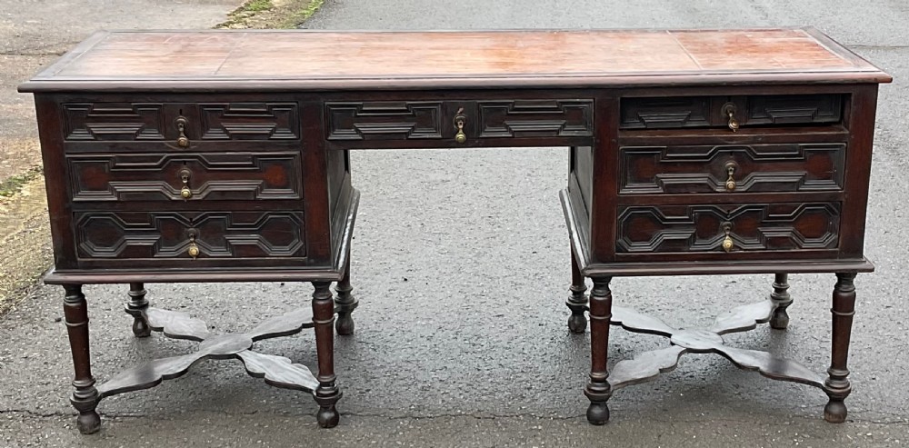 A large dark oak pedestal writing desk featuring geometric panelled drawer fronts, a leather inset writing surface, turned legs, and cross stretchers. View VI