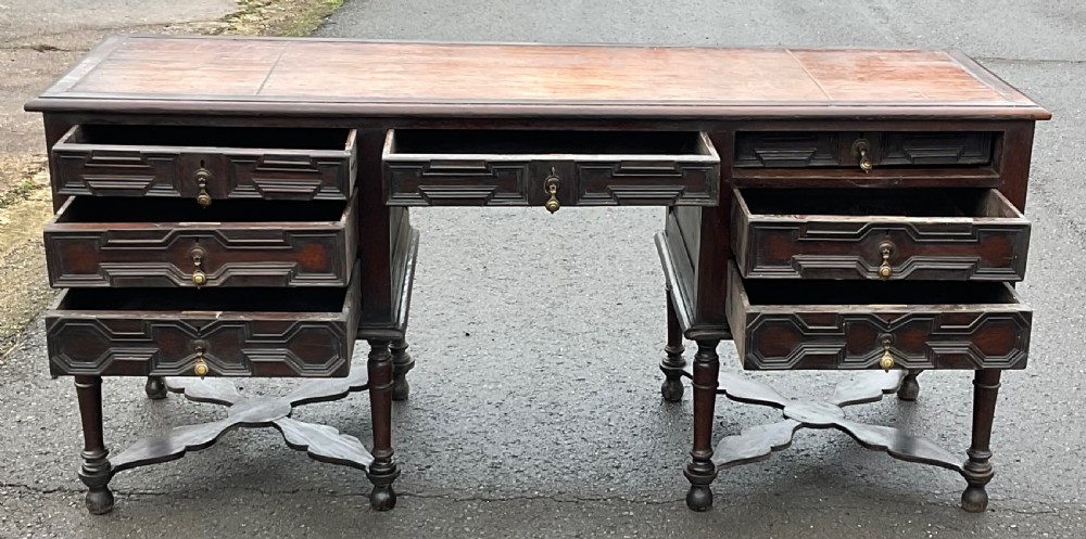 A large dark oak pedestal writing desk featuring geometric panelled drawer fronts, a leather inset writing surface, turned legs, and cross stretchers. View VII