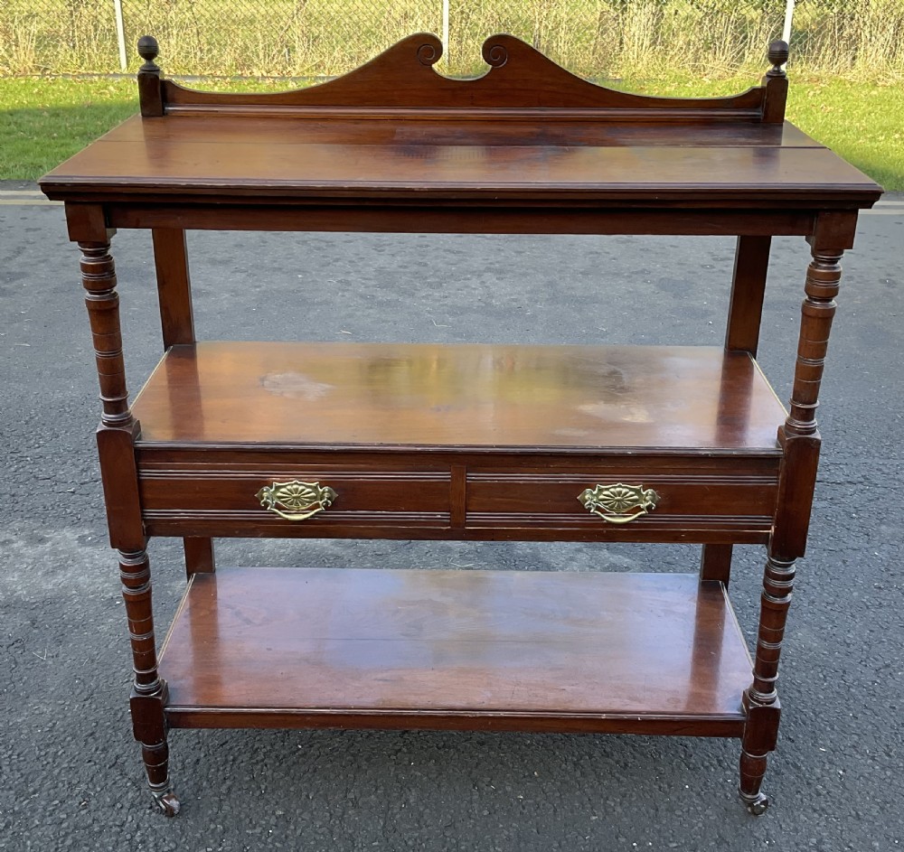 A Victorian mahogany three-tier stand featuring turned support posts, two drawers with decorative brass handles, a curved scroll top gallery, and brass castors.