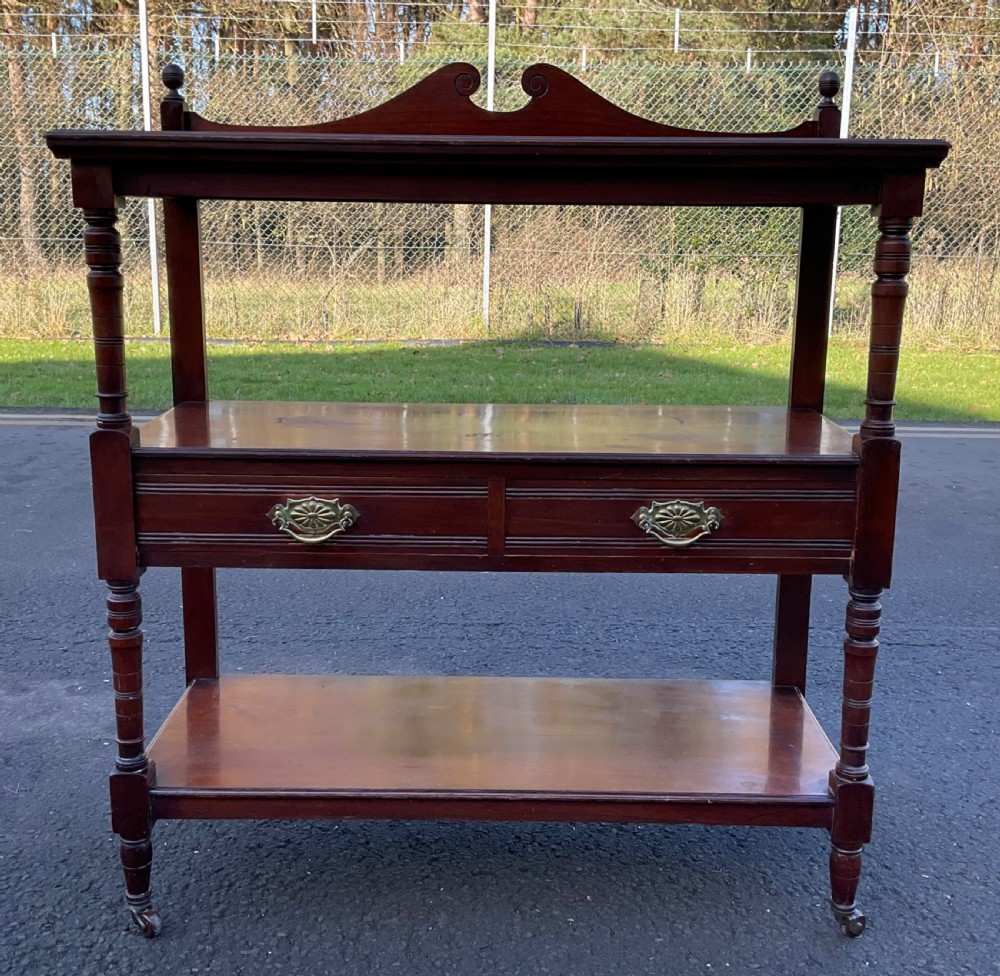 A Victorian mahogany three-tier stand featuring turned support posts, two drawers with decorative brass handles, a curved scroll top gallery, and brass castors. View II