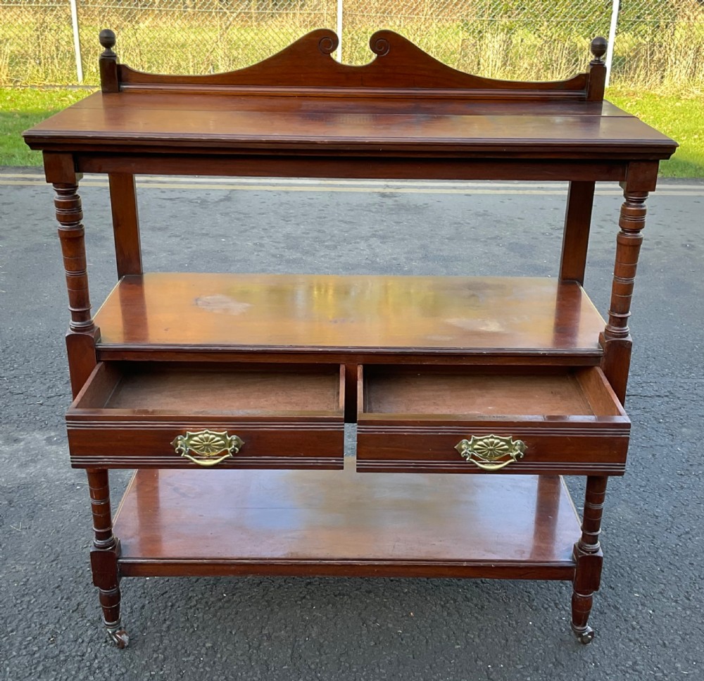 A Victorian mahogany three-tier stand featuring turned support posts, two drawers with decorative brass handles, a curved scroll top gallery, and brass castors. View V