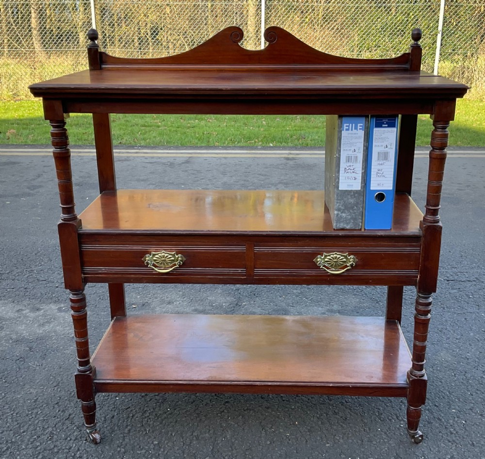 A Victorian mahogany three-tier stand featuring turned support posts, two drawers with decorative brass handles, a curved scroll top gallery, and brass castors. View VI