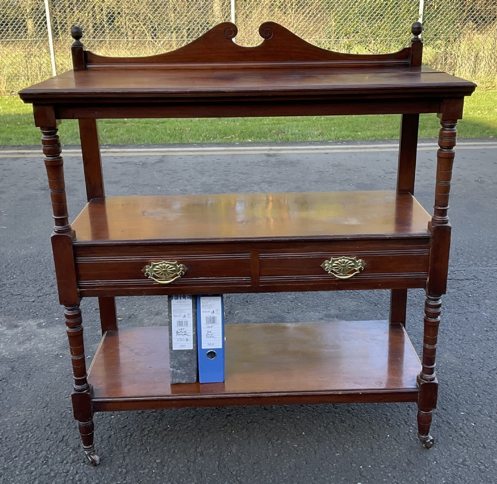 A Victorian mahogany three-tier stand featuring turned support posts, two drawers with decorative brass handles, a curved scroll top gallery, and brass castors. View VIII
