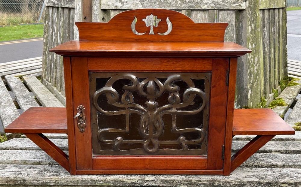 An Art Nouveau mahogany hanging wall cabinet featuring a central door with a decorative copper panel flanked by two side display shelves and topped with a pierced pediment.