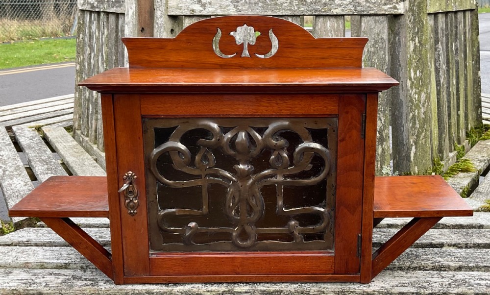 An Art Nouveau mahogany hanging wall cabinet featuring a central door with a decorative copper panel flanked by two side display shelves and topped with a pierced pediment. View II