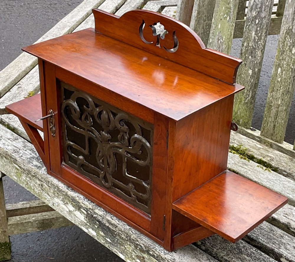 An Art Nouveau mahogany hanging wall cabinet featuring a central door with a decorative copper panel flanked by two side display shelves and topped with a pierced pediment. View V